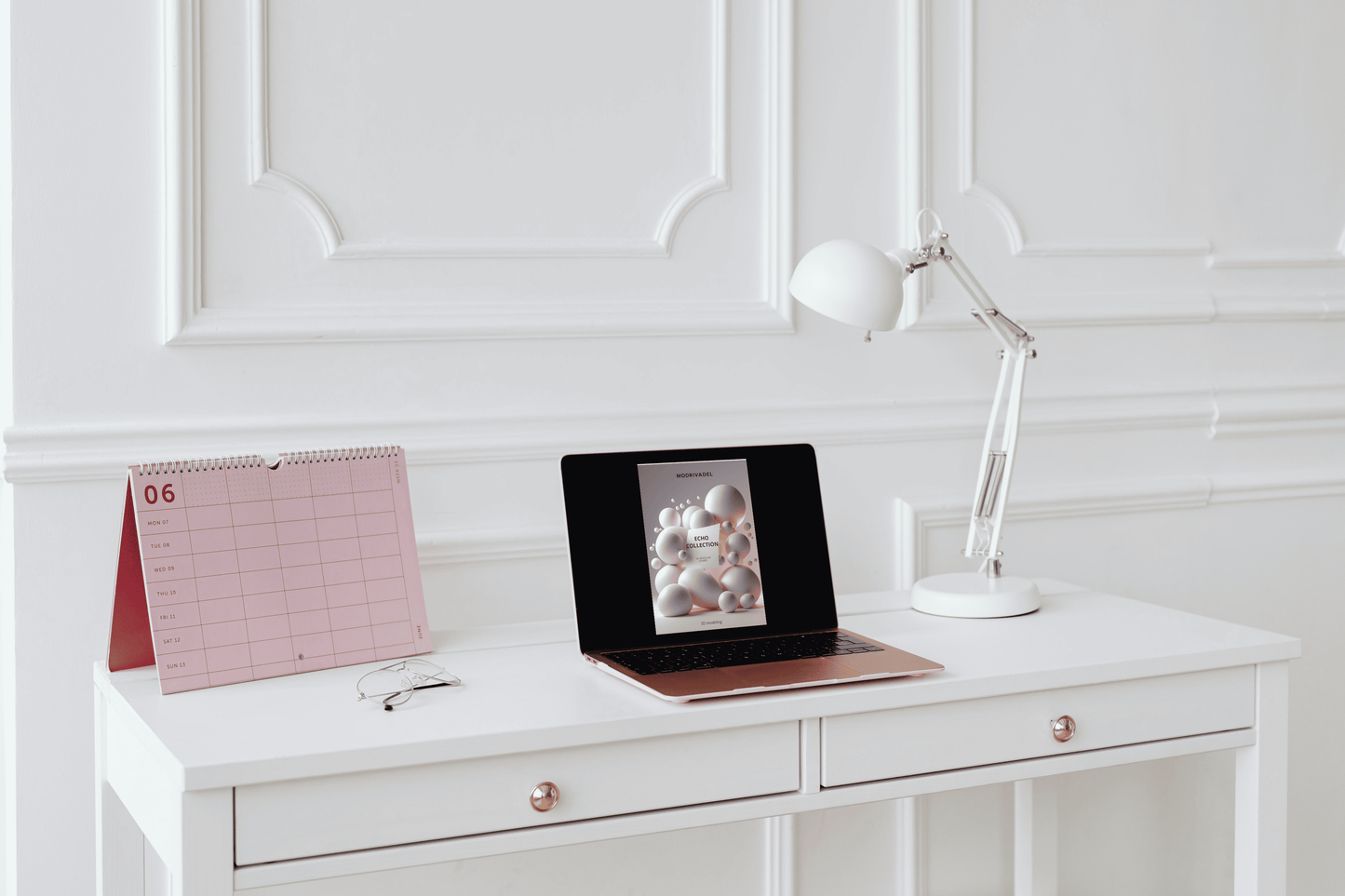 White desk with laptop, calendar, and lamp against a white paneled wall