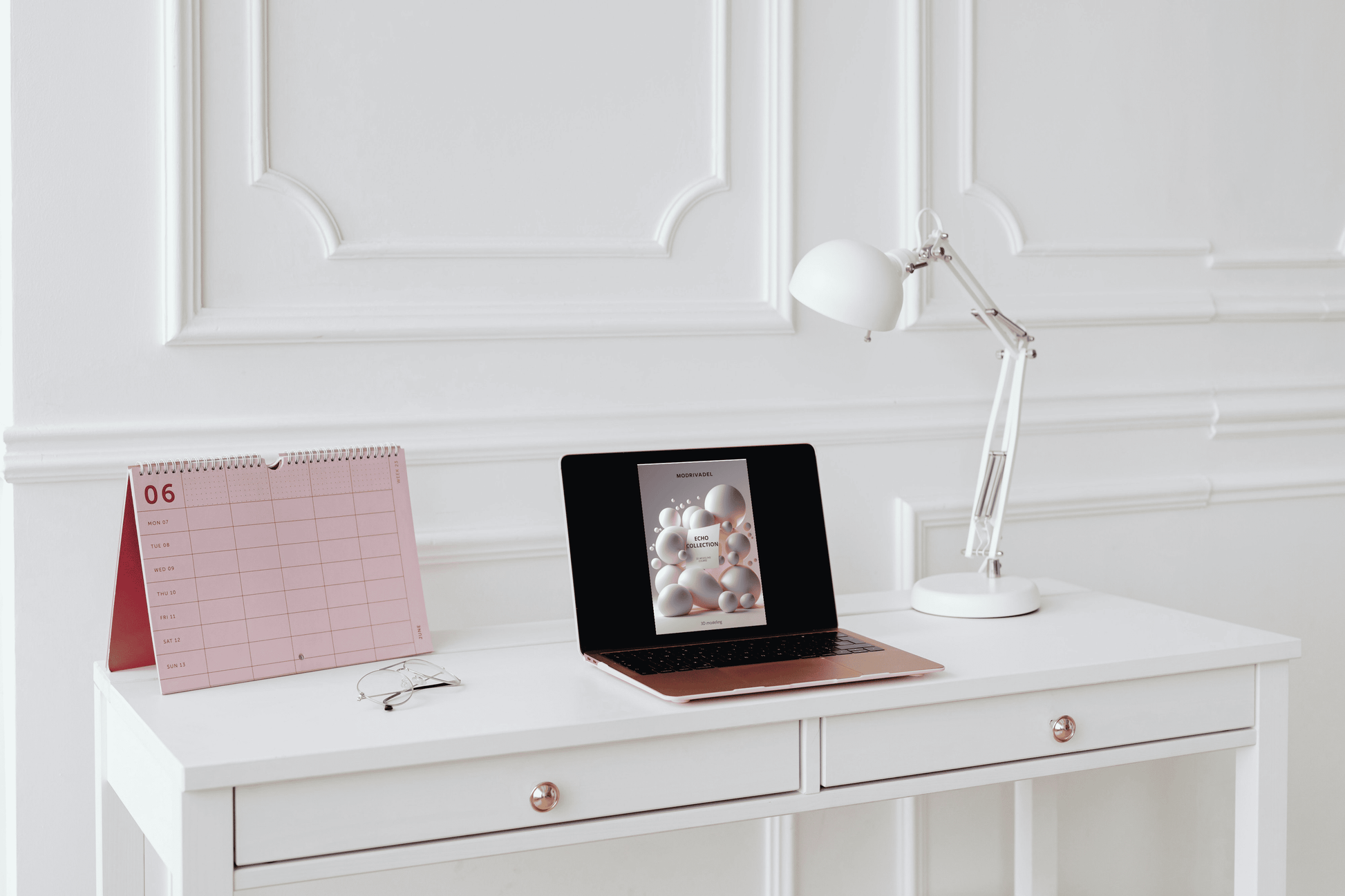 White desk with laptop, calendar, and lamp against a white paneled wall