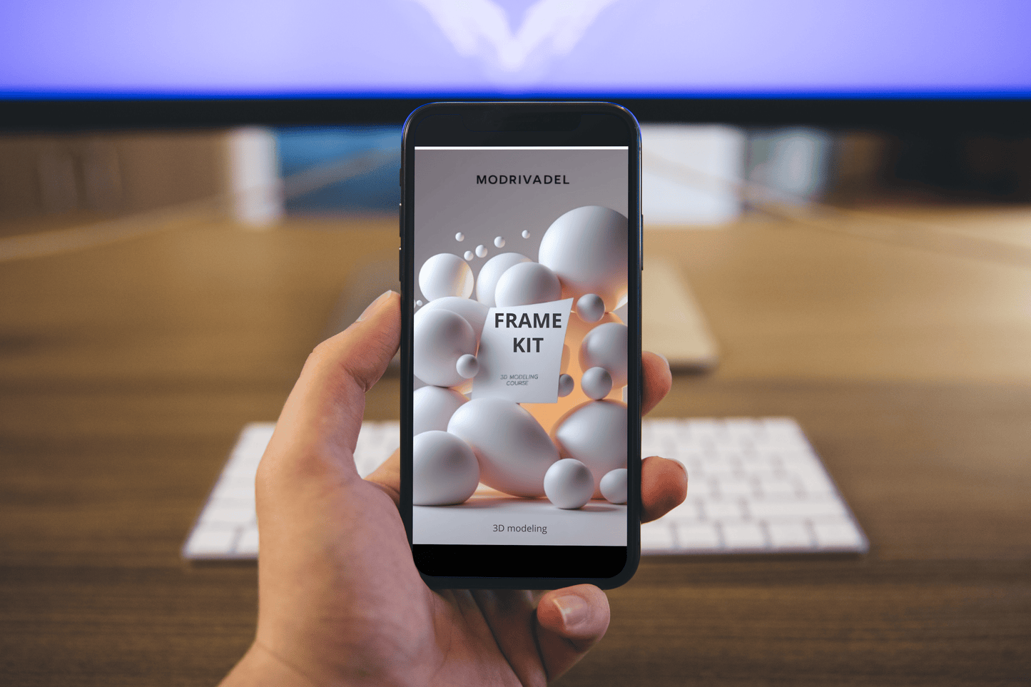 Hand holding a smartphone with a screen displaying 'MODRIVADEL FRAME KIT' on a blurred office desk background.