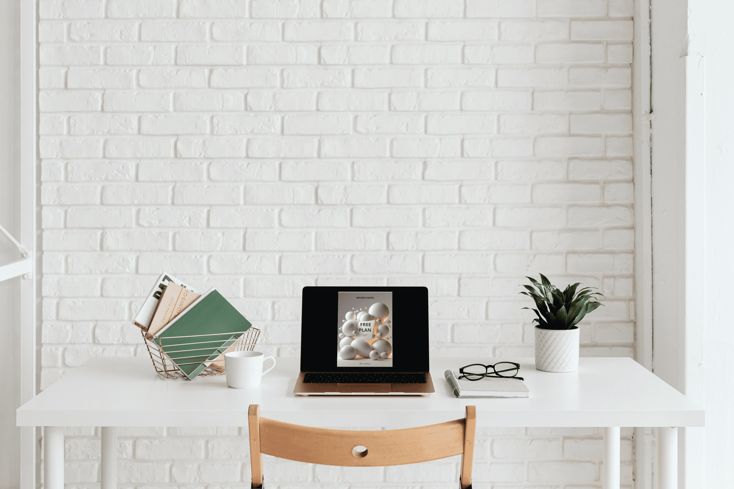 Minimalist desk setup with a laptop, mug, books, and plant against a white brick wall.