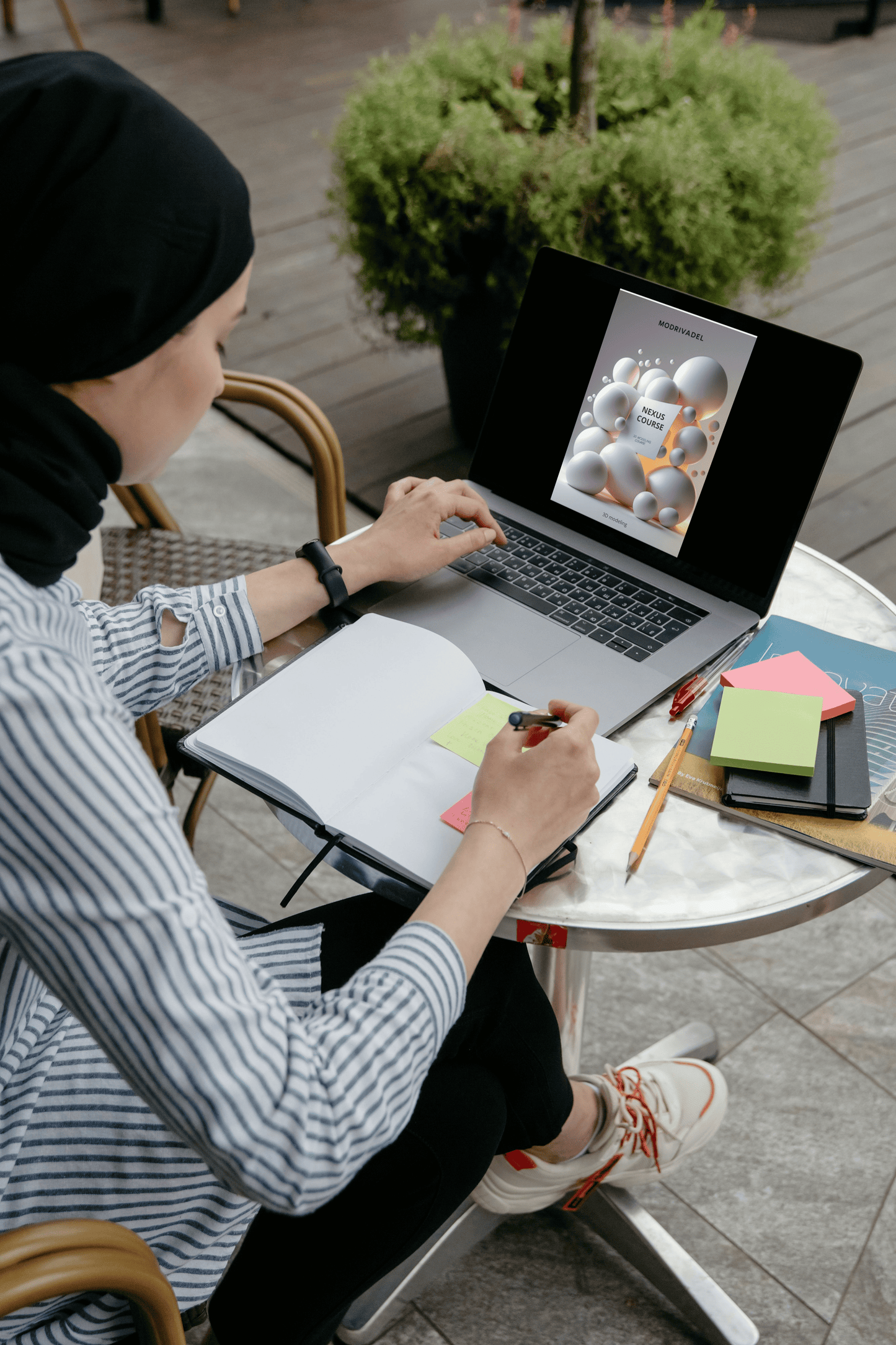 Person working on a laptop with a notebook and stationery at an outdoor table.