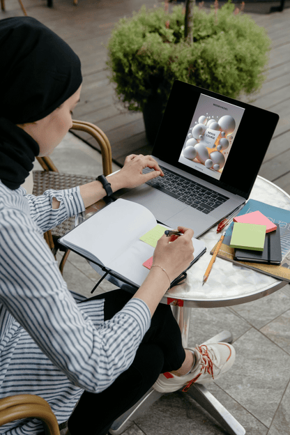 Person working on a laptop with a notebook and stationery at an outdoor table.