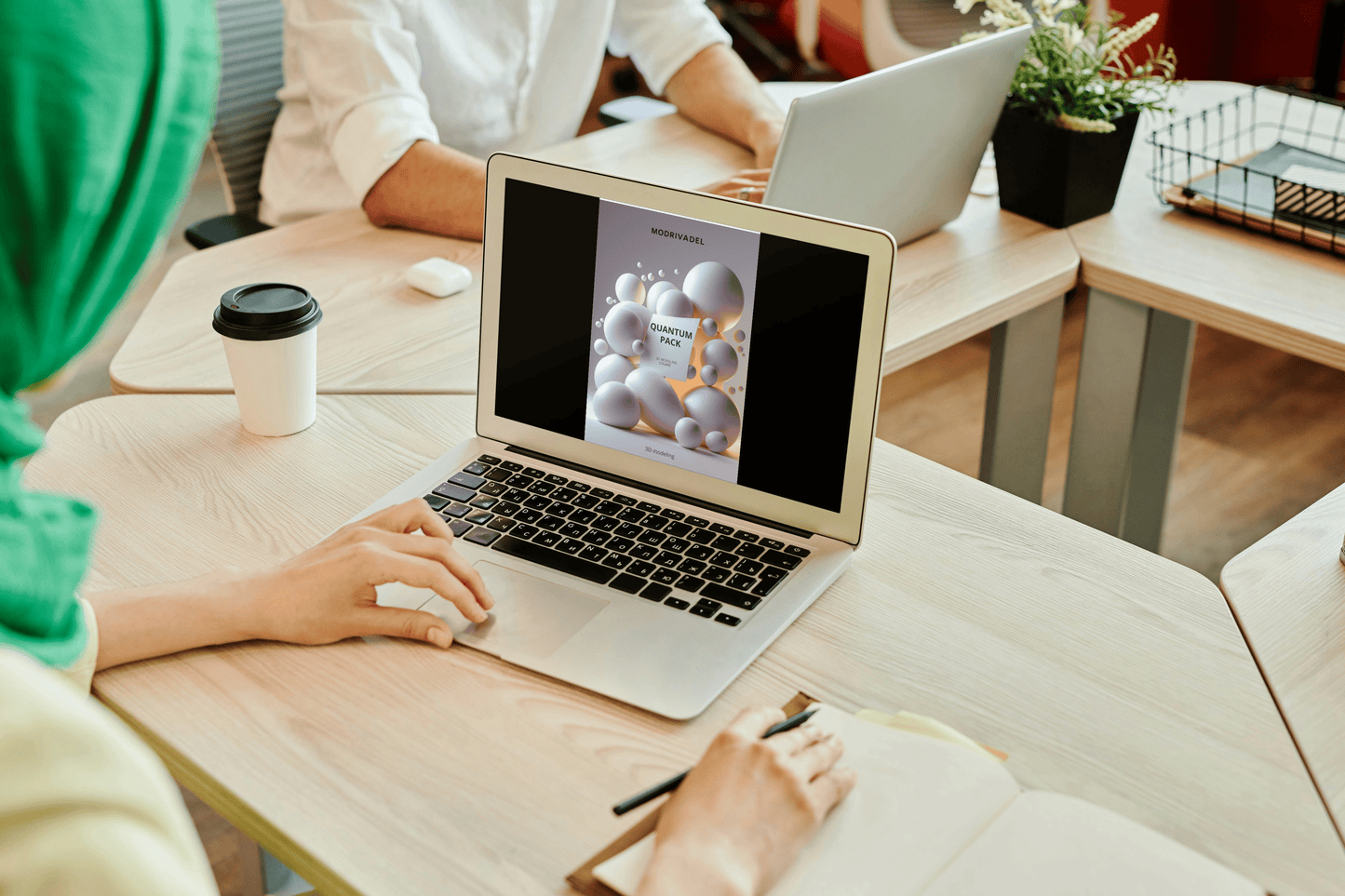 Person using a laptop with a colleague in the background at a desk.