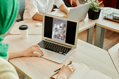 Person using a laptop with a colleague in the background at a desk.